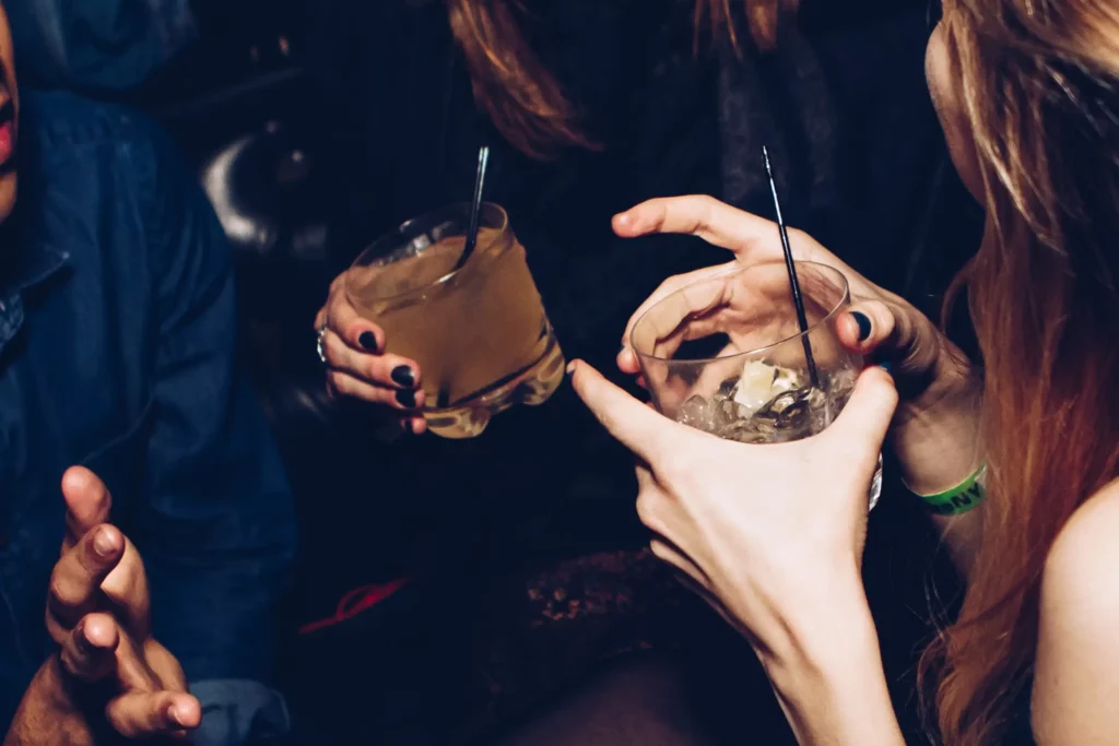 Close-up of two hands holding cocktails with straws in a social setting, with a dimly lit ambiance suggesting a bar or nightclub atmosphere. The focus on the drinks and hands implies a relaxed and enjoyable moment among friends