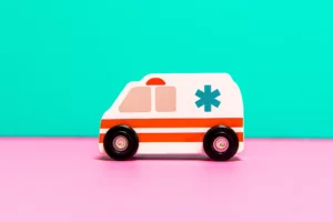 A wooden toy ambulance with simple, colorful details is placed against a vibrant pink and turquoise background. The toy features the iconic white body with red stripes and a blue star, representing an ambulance in a playful and child-friendly manner