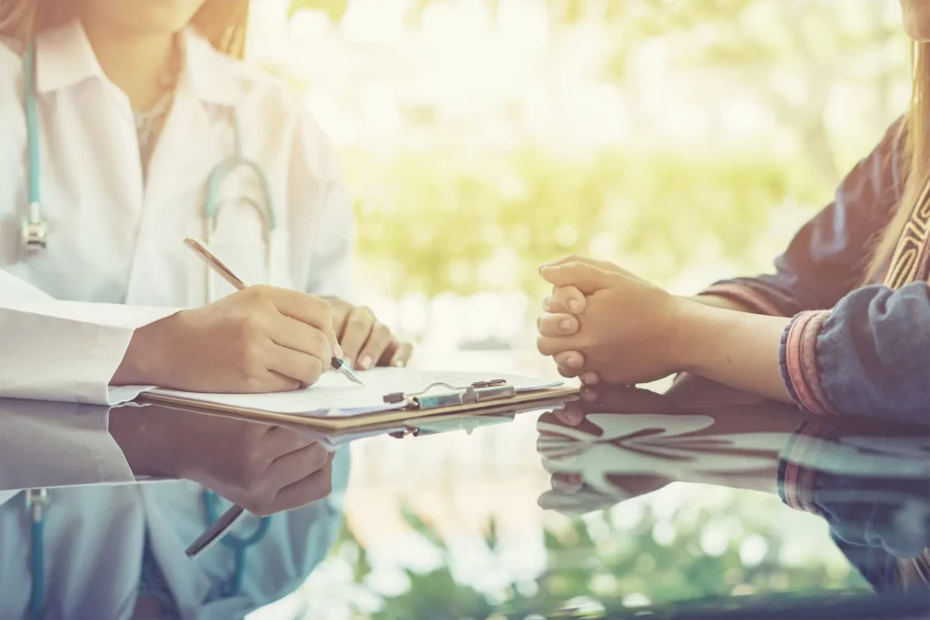 In a sunlit setting, a healthcare provider in a white coat is writing on a clipboard while conversing with a patient. The patient is sitting with hands clasped together, suggesting a dialogue or consultation is taking place. The bright, natural light and greenery in the background create a calming atmosphere for the medical consultation.