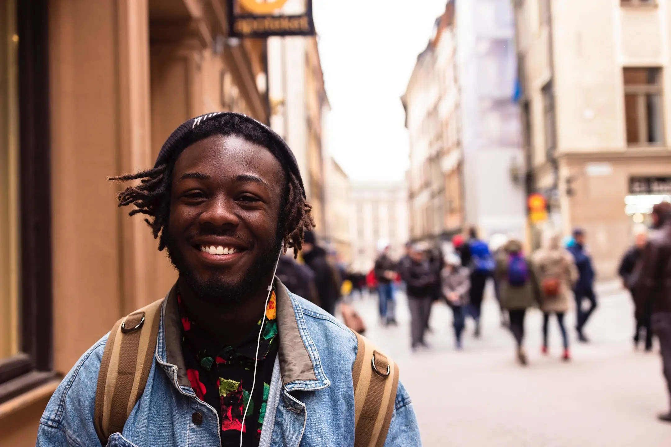 A joyful young man with dreadlocks wearing a beanie and denim jacket is smiling at the camera. He has earphones in and is carrying a messenger bag, suggesting he's enjoying music while out and about. The background is a busy city street scene with soft focus, highlighting the subject's happy demeanor in an urban setting