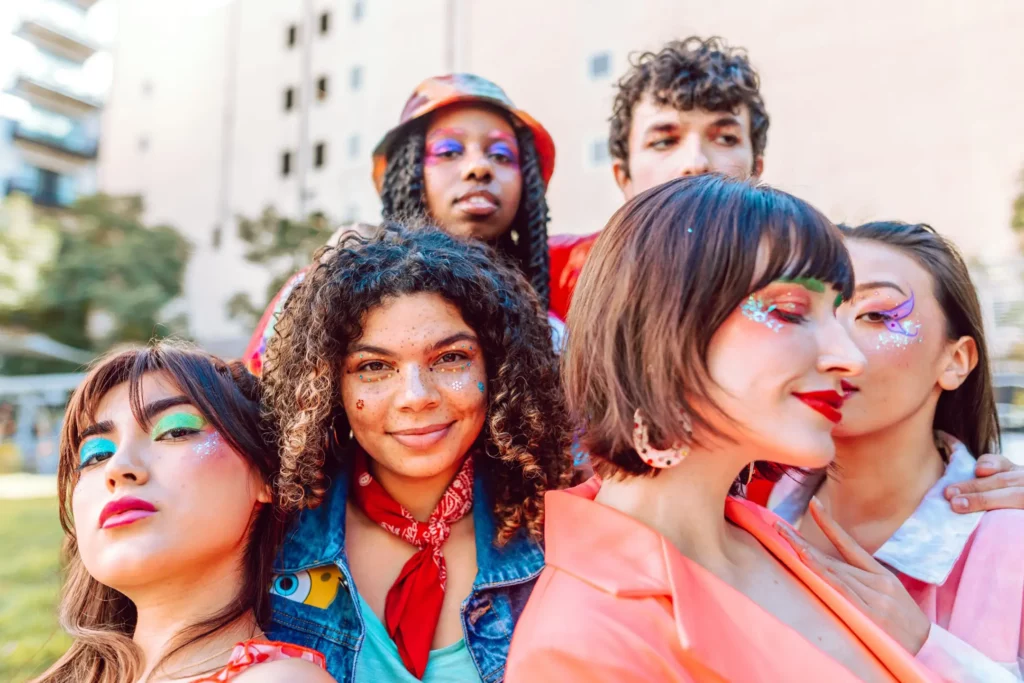 A diverse group of young adults with vibrant and artistic makeup are posing closely together. They each have unique styles, featuring colorful eyeshadow, glitter, and bold lipstick, and are wearing an assortment of fashionable outfits. Their expressions are happy and confident, reflecting a sense of friendship and creativity in an urban setting