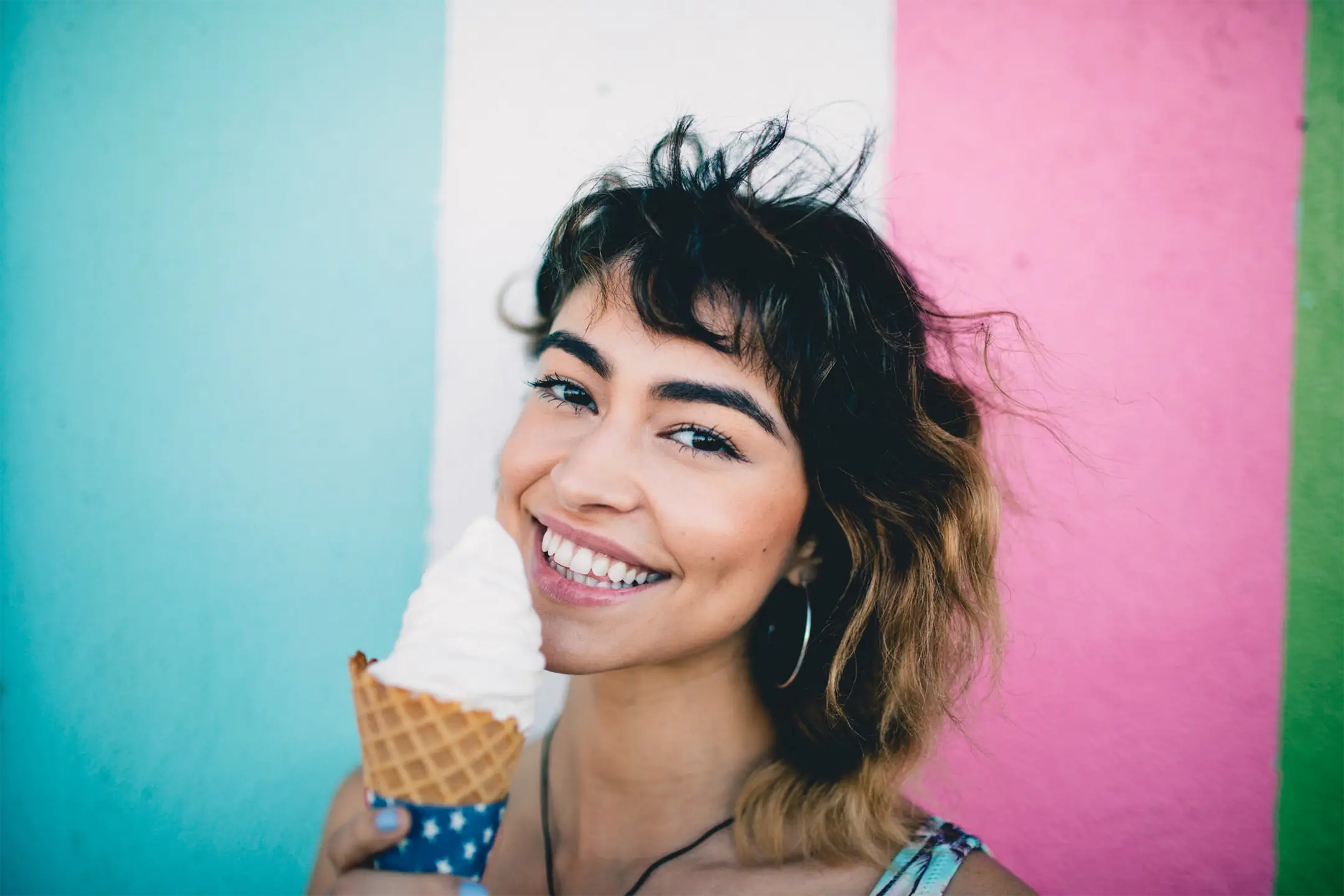 A joyful young woman with short curly brown hair, holding a vanilla ice cream cone in her hand, smiling brightly against a colorful background divided into three vertical stripes of turquoise, pink, and green