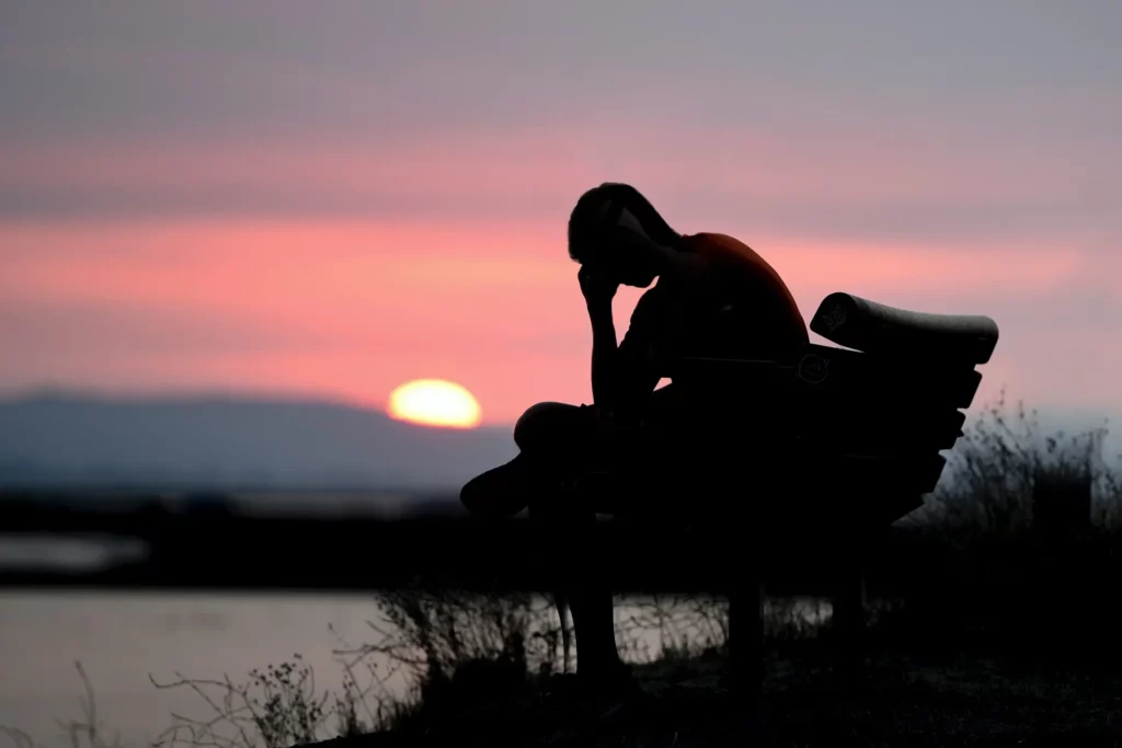 A silhouette of a person sitting on a bench, resting their head in their hand, against a backdrop of a stunning sunset with hues of pink and orange in the sky. The scene is tranquil, with the sun low on the horizon, indicating either dawn or dusk