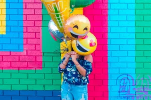 A person is obscured behind a cluster of vibrant helium balloons with emoji faces on them, against a multicolored brick wall backdrop. The balloons add a playful and joyous atmosphere to the scene, with the brightly colored wall enhancing the festive mood.