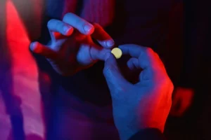 Illuminated by vibrant red and blue lights, a person's hands are shown holding a round pill between their fingers. The lighting casts a dramatic glow on the scene, highlighting the object and giving a mysterious ambiance to the action being performed.