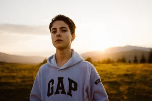 A young person with short hair stands in a field at sunset, wearing a hoodie with the word 'GAP' across the front. The golden hour light warmly illuminates their face and the background, featuring distant mountains and a soft sky, adding a serene and contemplative mood to the image.