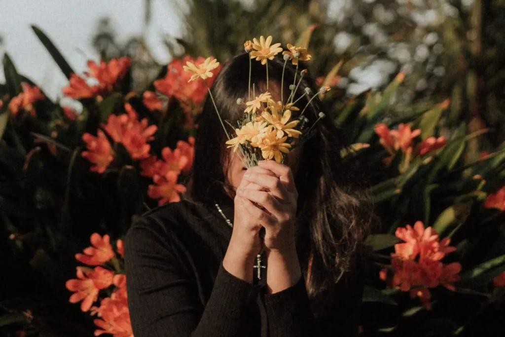 A person is holding a bouquet of yellow flowers in front of their face, obscuring it from view. They are standing against a lush backdrop of orange flowers and green foliage. The image has a dreamy quality, with the focus on the bouquet and the natural beauty surrounding the individual