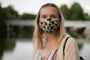 A young woman with blonde hair stands outdoors wearing a face mask with a sunflower print. She is looking directly at the camera with a calm and neutral expression, and the background is softly focused, featuring trees and a body of water, suggesting a tranquil outdoor setting