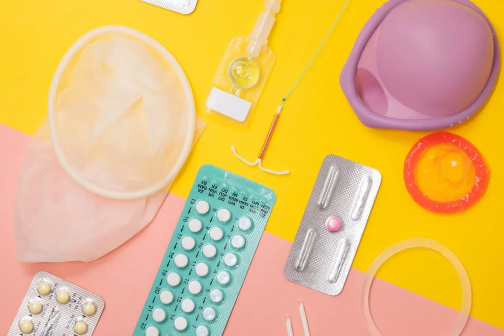 An assortment of contraceptive methods and sexual health products are arranged against a vibrant yellow background. Items include a diaphragm, condom, intrauterine device (IUD), contraceptive pills in blister packs, and emergency contraception pill, displaying a variety of options for birth control and protection.