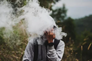 A person is obscured behind a thick cloud of smoke, with only their hand and a portion of their face partially visible. The smoke dominates the image, suggesting the act of smoking or vaping in an outdoor setting with foliage in the background, creating a sense of mystery or concealment.