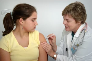 A young girl in a yellow shirt is receiving a vaccine injection in her upper arm from a medical professional, who appears to be a nurse or doctor. The healthcare worker is focused on the injection, ensuring proper technique, while the girl looks away, possibly to avoid the sight of the needle. Both are indoors, and the setting suggests a clinical environment conducive to medical procedures