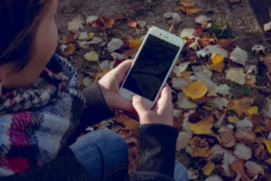 A person is seated outside amidst autumn leaves, focusing on a smartphone held in their hands, with their face obscured and attention on the device, blending technology use with the natural backdrop of fall
