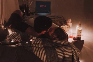 A couple is lying down in a cozy embrace, illuminated by the soft glow of string lights, with a laptop showing Netflix in the background. A romantic setting is enhanced by a bottle of wine, a glass, and candles, creating an intimate atmosphere for a movie night.