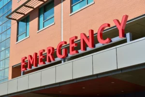 The bold red lettering of the word 'EMERGENCY' is prominently displayed on the facade of a modern hospital building. The letters are mounted on an overhang above the entrance, with a clear blue sky in the background and part of the building's brick and glass exterior visible