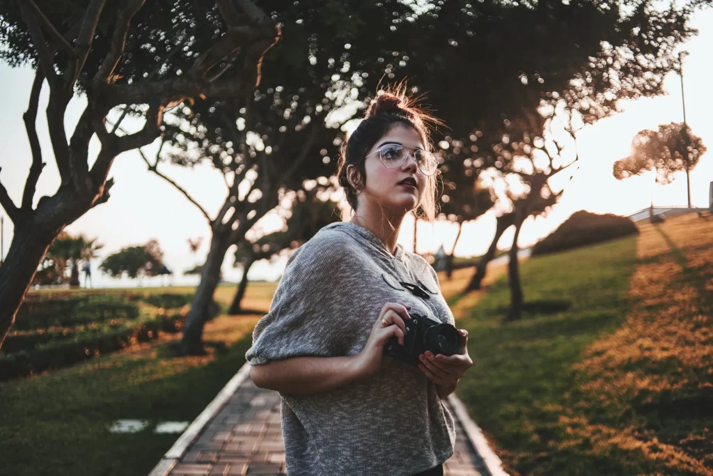 A young woman with glasses and her hair up is holding a camera, standing on a wooden path in a park at sunset. The warm light of the setting sun highlights her profile and the surrounding trees, creating a serene atmosphere. Her thoughtful expression suggests she is searching for the perfect shot or reflecting on a recent capture